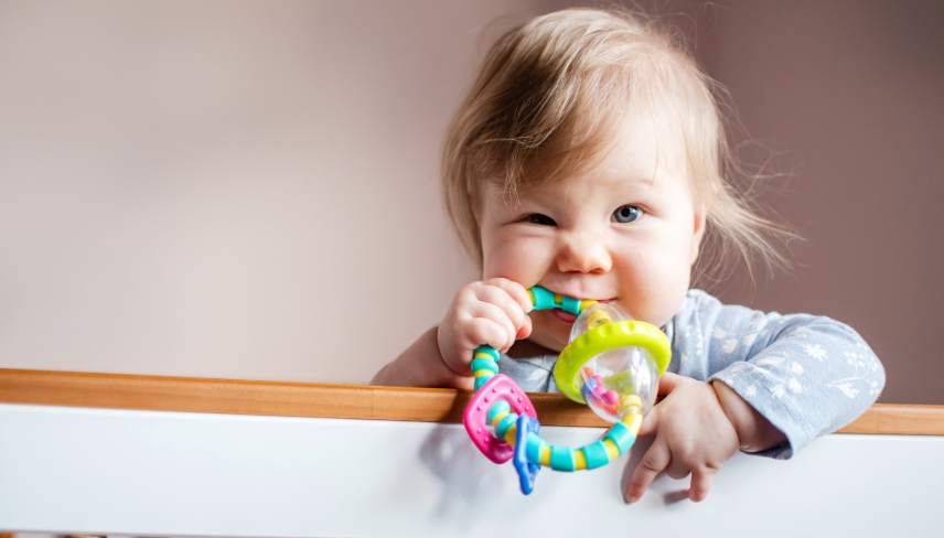 Cute Caucasian little baby girl standing in her crib and chewing on an infant toy, looking at the camera, with copy space. 