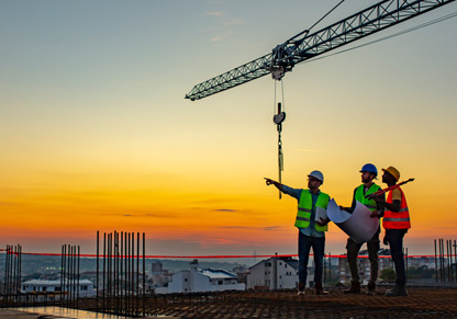 Three people stand on top of a building wearing safety gear, with a crane and an orange sky in the background. 