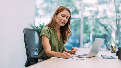 A woman sitting at a desk, writing in a notebook while using a laptop in a bright office setting.